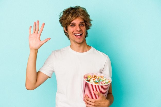 Jonge blanke man met make-up met popcorn geïsoleerd op blauwe achtergrond glimlachend vrolijk nummer vijf met vingers.