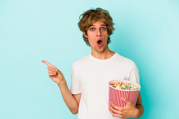 Jonge blanke man met make-up met popcorn geïsoleerd op een blauwe achtergrond die naar de zijkant wijst