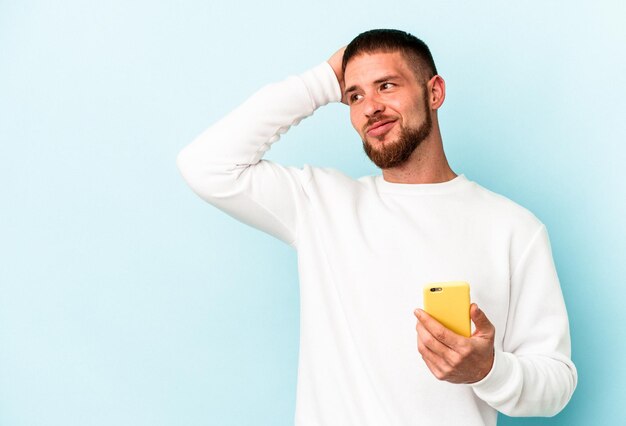 Jonge blanke man met mobiele telefoon geïsoleerd op blauwe achtergrond achterhoofd aanraken, denken en een keuze maken.