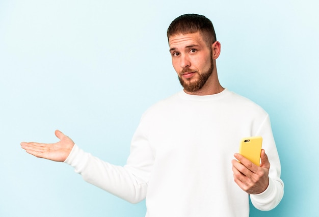 Jonge blanke man met mobiele telefoon geïsoleerd op blauwe achtergrond met een kopie ruimte op een palm en met een andere hand op de taille.
