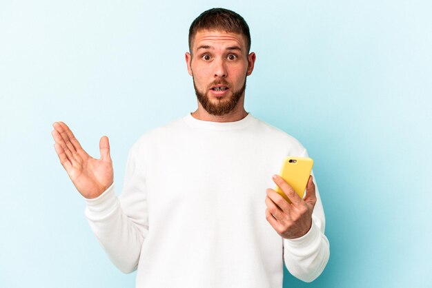 Jonge blanke man met mobiele telefoon geïsoleerd op blauwe achtergrond verrast en geschokt.