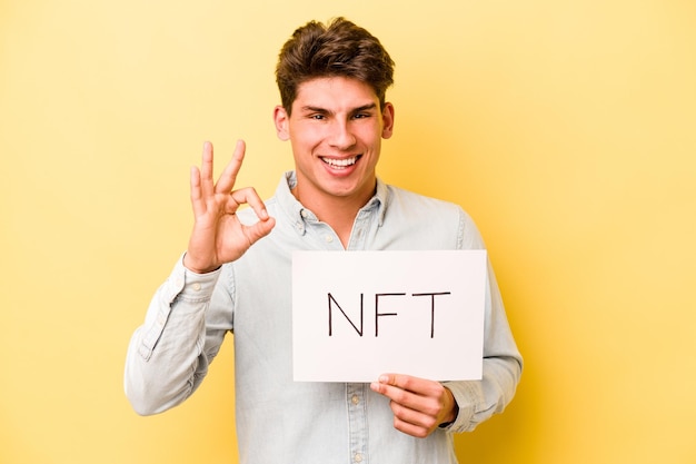 Jonge blanke man met NFT plakkaat geïsoleerd op gele achtergrond vrolijk en zelfverzekerd met ok gebaar