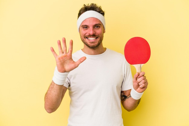 Jonge blanke man met pingpongracket geïsoleerd op gele achtergrond glimlachend vrolijk nummer vijf met vingers.