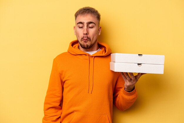 Jonge blanke man met pizza's geïsoleerd op gele achtergrond haalt zijn schouders op en open ogen verward.