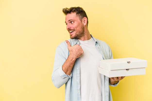 Jonge blanke man met pizza's geïsoleerd op gele achtergrond wijst met duimvinger weg, lachend en zorgeloos.