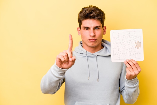 Jonge blanke man met puzzel geïsoleerd op gele achtergrond met nummer één met vinger