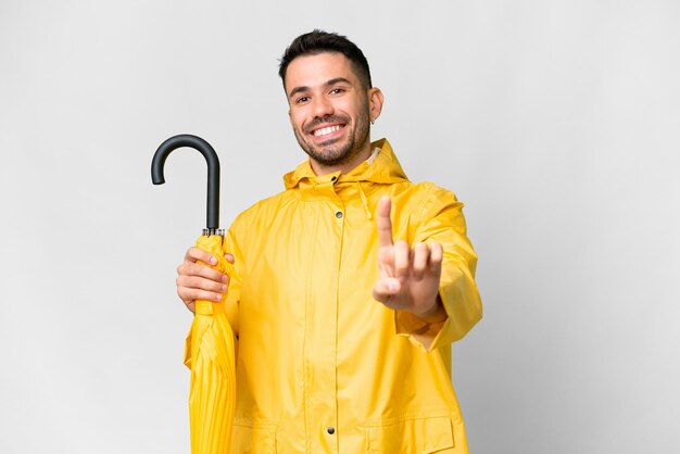Jonge blanke man met regenbestendige jas en paraplu over geïsoleerde witte achtergrond die een vinger laat zien en optilt