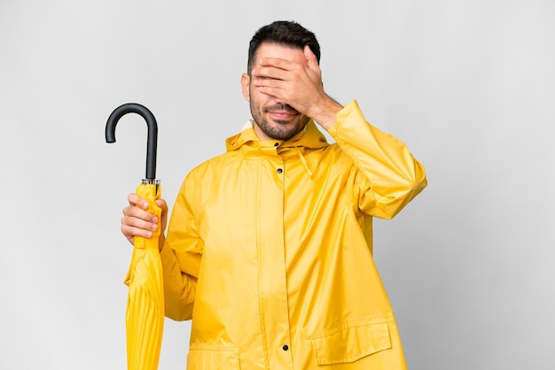 Jonge blanke man met regendichte jas en paraplu over geïsoleerde witte achtergrond die de ogen met de handen bedekt ik wil niets zien