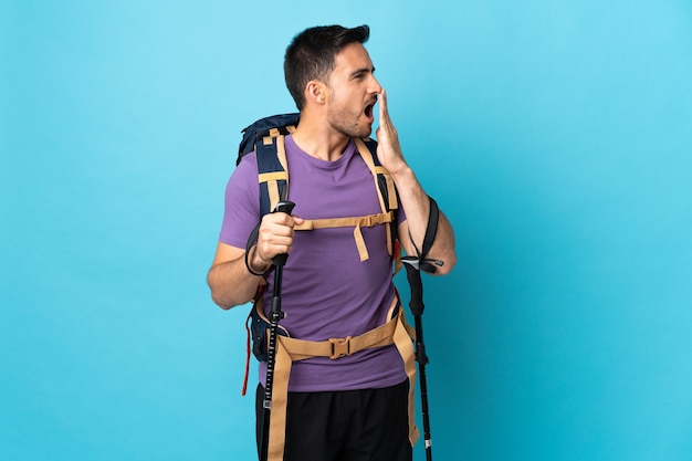 Jonge blanke man met rugzak en wandelstokken geïsoleerd op blauw geeuwen en die wijd open mond met de hand bedekken