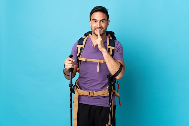 Jonge blanke man met rugzak en wandelstokken geïsoleerd op blauw met een teken van stilte gebaar vinger in de mond steken