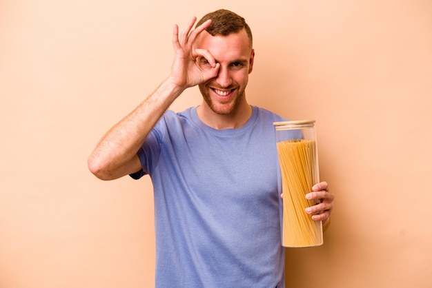 Jonge blanke man met spaghetti pot geïsoleerd op beige achtergrond opgewonden houden ok gebaar in de gaten