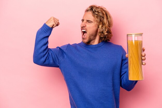 Jonge blanke man met spaghetti pot geïsoleerd op roze achtergrond die vuist opheft na een overwinning, winnaar concept.