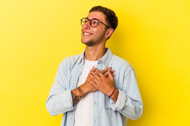 Jonge blanke man met tatoeages geïsoleerd op gele achtergrond lachen houden handen op het hart, concept van geluk.