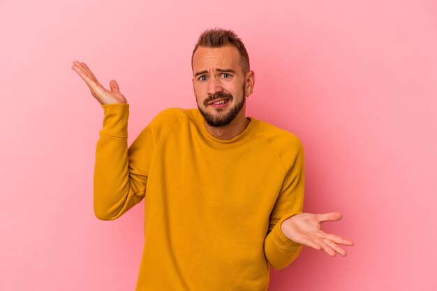 Jonge blanke man met tatoeages geïsoleerd op roze achtergrond verward en twijfelachtig schouders ophalen om een kopie ruimte te houden.