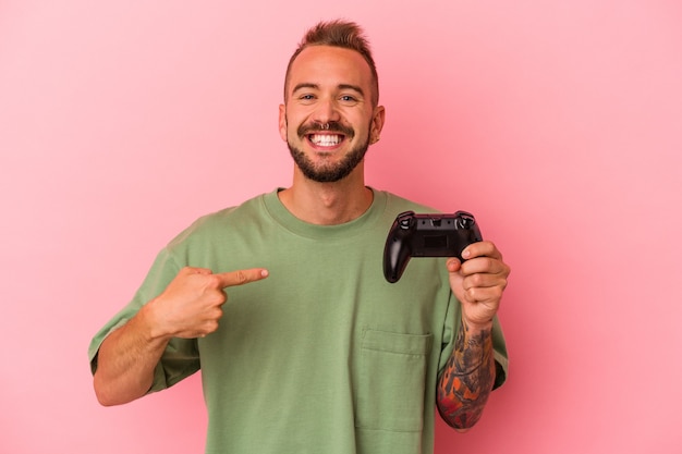 Jonge blanke man met tatoeages met gamecontroller geïsoleerd op een roze achtergrond persoon die met de hand wijst naar een shirt kopieerruimte, trots en zelfverzekerd?