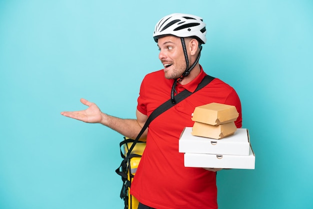 Jonge blanke man met thermische rugzak en fastfood geïsoleerd op blauwe achtergrond met verbaasde uitdrukking terwijl hij naar de zijkant kijkt