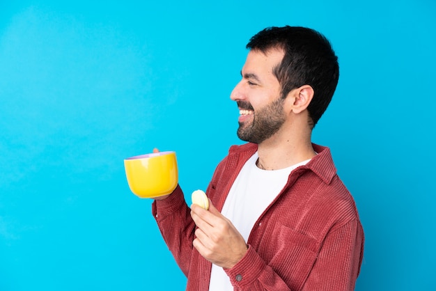 Jonge blanke man over geïsoleerde blauwe muur met kleurrijke Franse macarons en een kopje melk