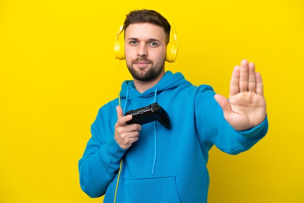 Jonge blanke man speelt met een videogamecontroller geïsoleerd op een gele achtergrond die een stopgebaar maakt