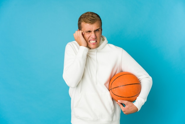 Jonge blanke man spelen basketbal geïsoleerd op blauwe achtergrond voor oren met handen.