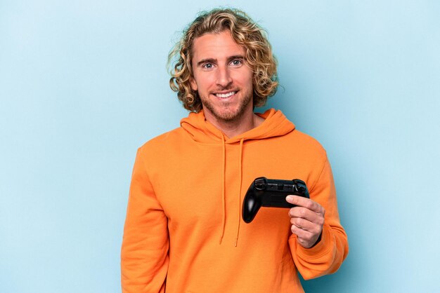 Jonge blanke man spelen met een video game controller geïsoleerd op blauwe achtergrond gelukkig, lachend en vrolijk.