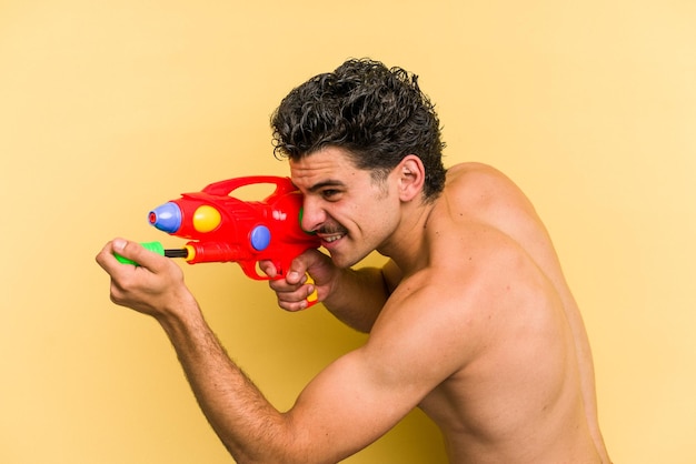 Jonge blanke man spelen met een waterpistool geïsoleerd op gele achtergrond