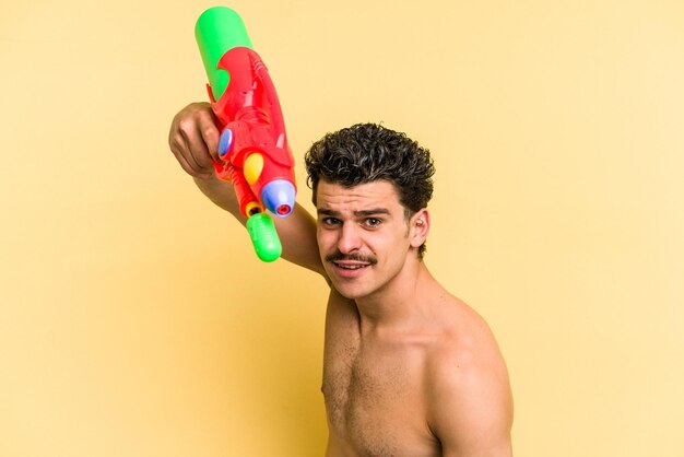 Jonge blanke man spelen met een waterpistool geïsoleerd op gele achtergrond