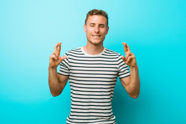 Jonge blanke man tegen een blauwe muur kruising vingers voor het hebben van geluk