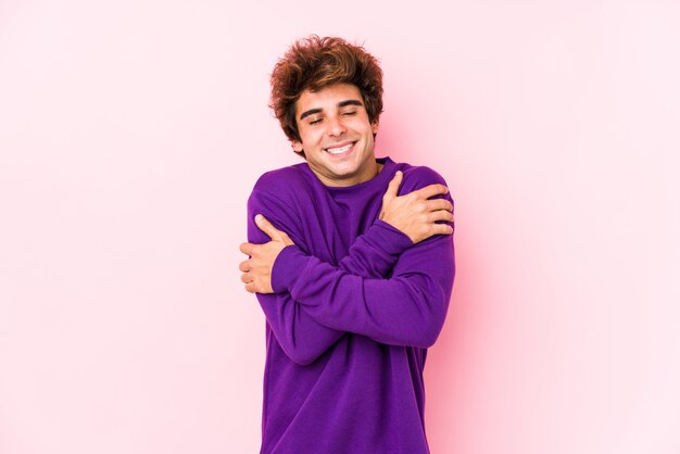 Jonge blanke man tegen een roze muur knuffels, glimlachend zorgeloos en gelukkig.