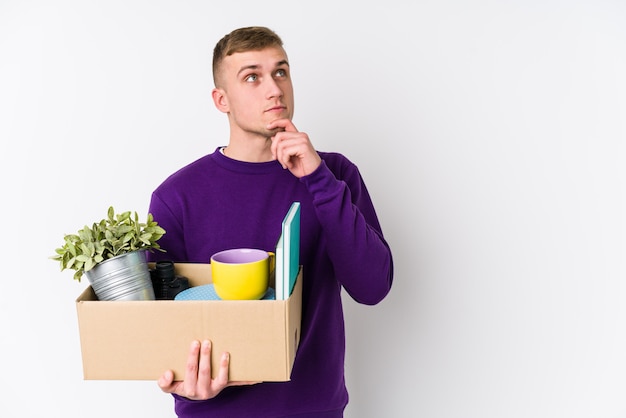 Jonge blanke man verhuizen naar een nieuw huis zijwaarts op zoek met twijfelachtige en sceptische uitdrukking.