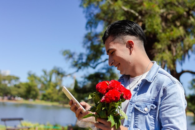 Jonge blanke man verliefd op bloemen in de hand die een bericht verzendt met zijn telefoon buiten in een openbaar park met een natuurlijke habitat, een lagune en bomen op de achtergrond