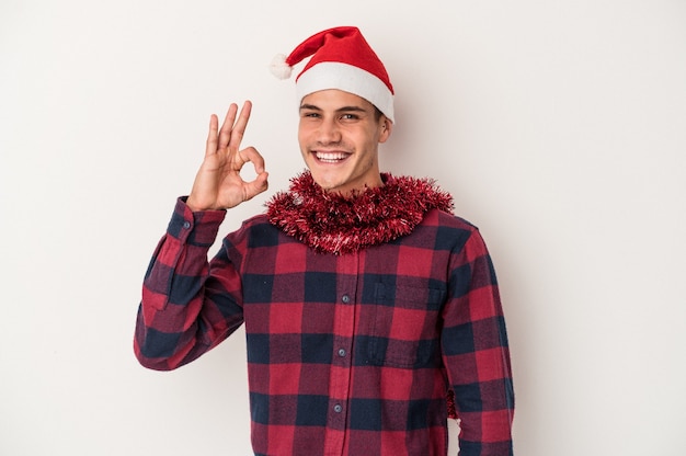 Jonge blanke man viert Kerstmis geïsoleerd op een witte achtergrond vrolijk en zelfverzekerd weergegeven: ok gebaar.