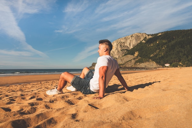 Jonge blanke man zit op het zand van een Baskisch strand.