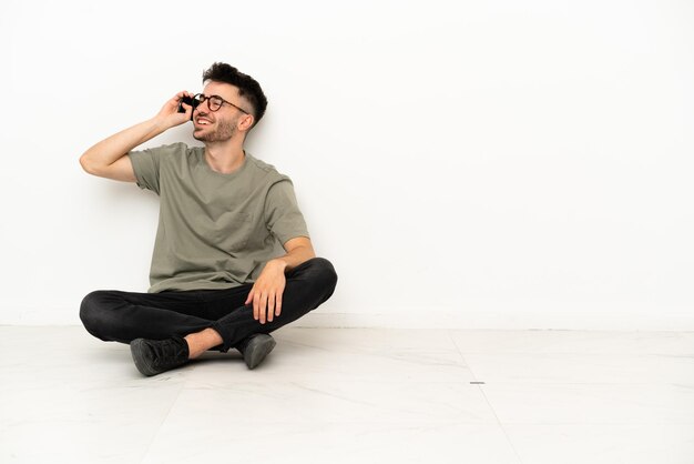 Jonge blanke man zittend op de vloer geïsoleerd op een witte achtergrond een gesprek voeren met de mobiele telefoon