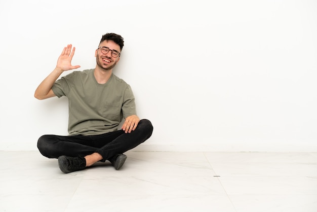 Jonge blanke man zittend op de vloer geïsoleerd op een witte achtergrond saluerend met de hand met gelukkige uitdrukking
