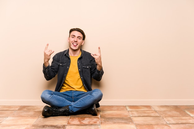 Jonge blanke man zittend op de vloer met rock gebaar met vingers