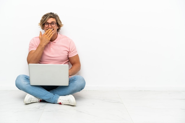 Jonge blanke man zittend op de vloer met zijn laptop geïsoleerd op een witte achtergrond gelukkig en lachend die mond bedekt met hand