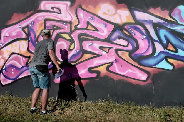 Jonge blanke mannelijke graffitikunstenaar die grote straatkunst schildert in blauwe en roze tinten