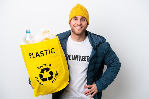 Jonge blanke met een zak vol plastic flessen om te recyclen geïsoleerd op een witte achtergrond poseren met armen op de heup en glimlachen