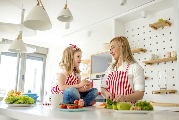 Jonge blanke moeder in gesprek met haar dochter op het aanrecht, het dragen van bijpassende schorten in een keuken