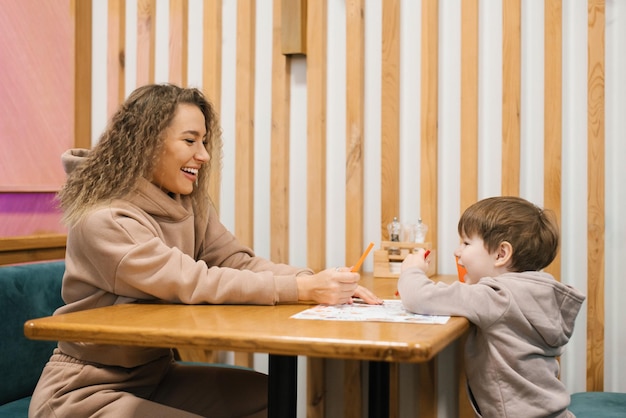 Jonge blanke moeder zit aan een tafel in een café met haar vier jaar oude zoon te wachten op haar bestelling en plezier te maken met het communiceren met elkaar
