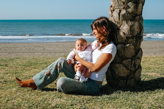 Jonge blanke moeder zittend op het gras met baby in haar armen aan zee