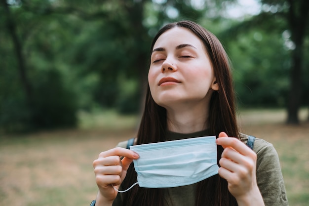 Jonge blanke mooie vrouw verwijdert een medisch beschermend masker op de natuur, schone frisse lucht inademen.