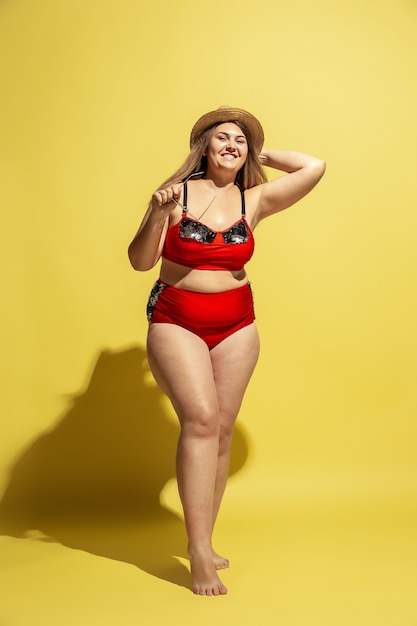 Jonge blanke plus size vrouwelijk model bereidt zich voor op strandresort op gele muur. Vrouw in rode zwembroek, hoed en zonnebril. Concept van zomer, feest, lichaam positief, gelijkheid en chill.
