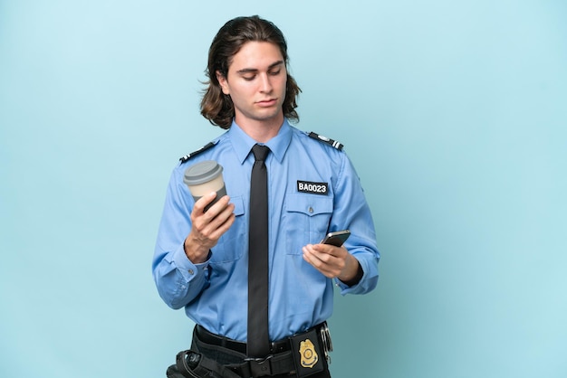 Jonge blanke politieman geïsoleerd op blauwe achtergrond met koffie om mee te nemen en een mobiel