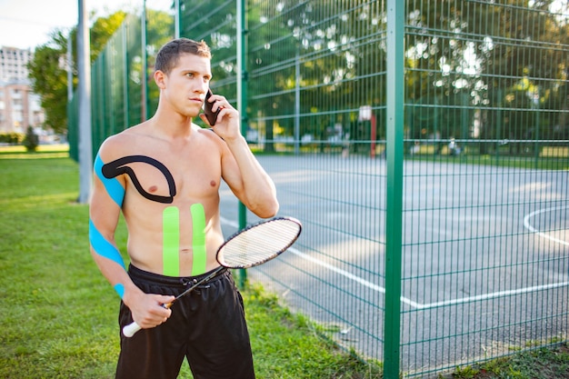 Jonge blanke professionele tennisser met kinesiologie taping op lichaam dragende racket in de buurt van omheind sportveld