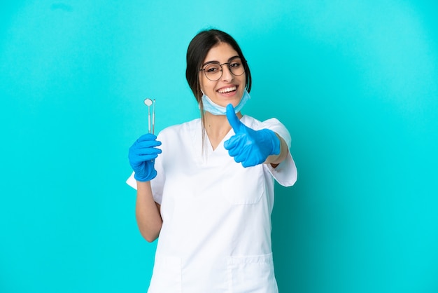 Jonge blanke tandartsvrouw met gereedschap geïsoleerd op een blauwe achtergrond met duimen omhoog omdat er iets goeds is gebeurd