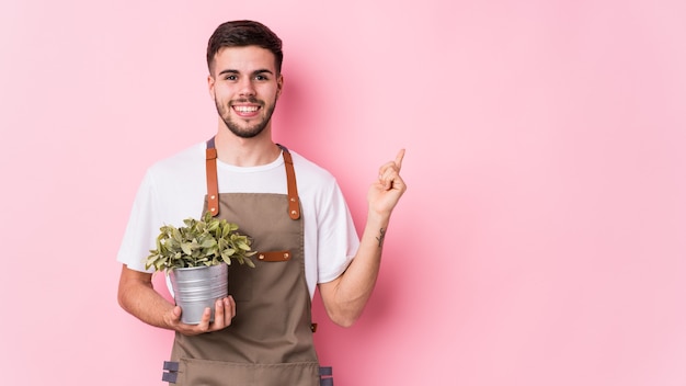 Jonge blanke tuinman man met een plant geïsoleerd glimlachend en opzij wijzend, iets laten zien op lege ruimte.