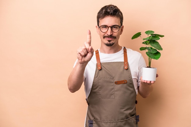 Jonge blanke tuinman man met een plant geïsoleerd op een beige achtergrond met nummer één met vinger