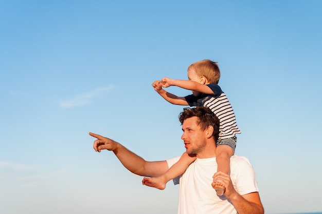 Jonge blanke vader met zoontje lopen warme zomerdag langs de kust. Zomer familievakantie concept. Vriendschap vader en zoon. De zoon van de vaderholding op zijn schouders bij het strand