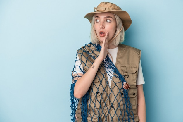 Jonge blanke vissersvrouw die een net vasthoudt dat op een blauwe achtergrond is geïsoleerd, zegt een geheim heet remnieuws en kijkt opzij